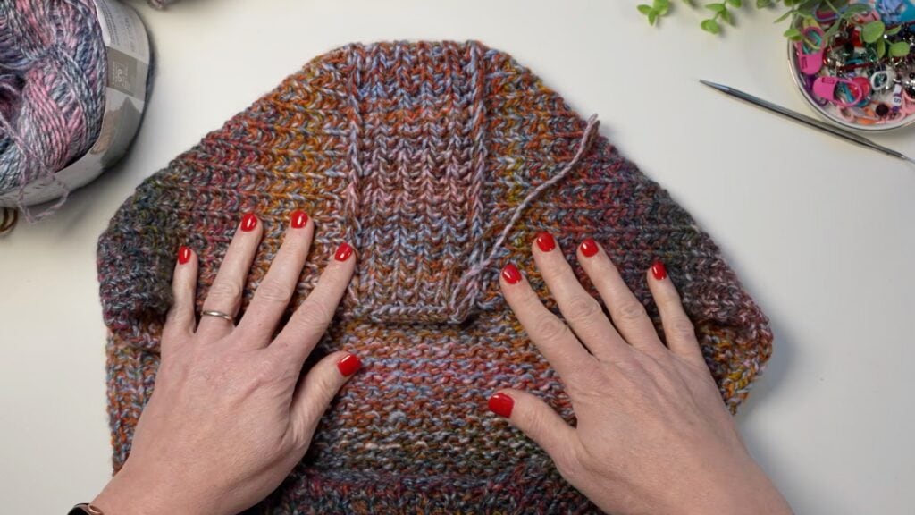 Hands with red nails working on a vibrant knit piece, with visible stitching, yarn, needles, buttons, and some green leaves nearby.