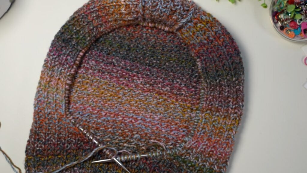 A multicolored circular knitting project in progress on metal needles, with visible stitch detail, rests among crafting supplies.