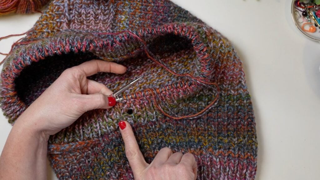 Hands with red nail polish knit a vibrant, textured piece with silver needles, highlighting a stitch detail on a white table.