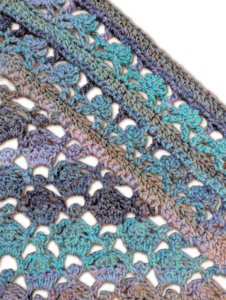 Diagonal crocheted fabric with an open shell stitch, using variegated blue, green, purple, and brown yarn for a lacy texture.