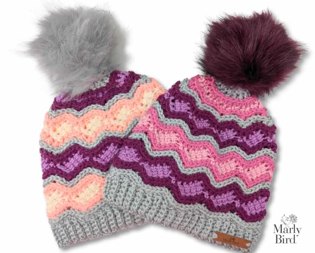 Perfect for those First Fall Weekends, these knitted hats showcase wavy patterns and playful pom-poms. The left hat blends pastel pink, peach, purple, and gray with a gray pom-pom, while the right features pink, purple, and gray tones topped with a dark brown pom-pom. Both sport ribbed brims. -Marly Bird