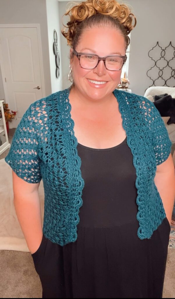 A person with glasses and a warm smile stands indoors, wearing a black top and a Garden Party Crochet Cardigan. They have their hands in their pockets. The background shows home decor, including a door, wall art, and part of a couch with a throw blanket. -Marly Bird