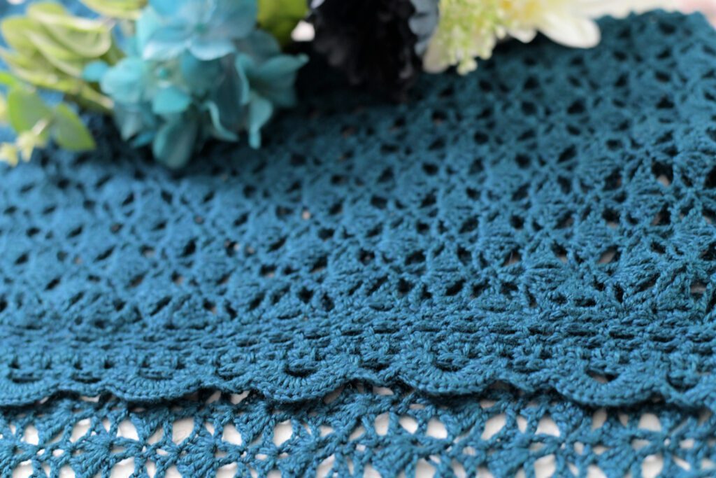A close-up image of a crocheted scalloped edge in a teal fabric with intricate patterns, reminiscent of a cozy crochet cardigan. In the background, partly blurred, there are a few flowers with light blue, white, and green petals, adding a touch of color to the scene. Garden Party Crochet Cardigan Pattern-Marly Bird
