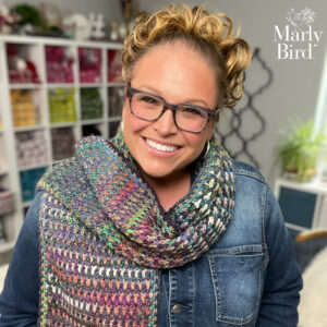 Woman models a vibrant crochet scarf with textured stitches, styled over a denim jacket; yarn shelves and plant behind her.