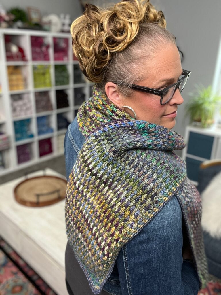 A woman with curly blonde hair in an updo wears glasses, a denim jacket, and a multi-colored, textured knit shawl. She stands in a cozy room with yarn cubes in the background. -Marly Bird