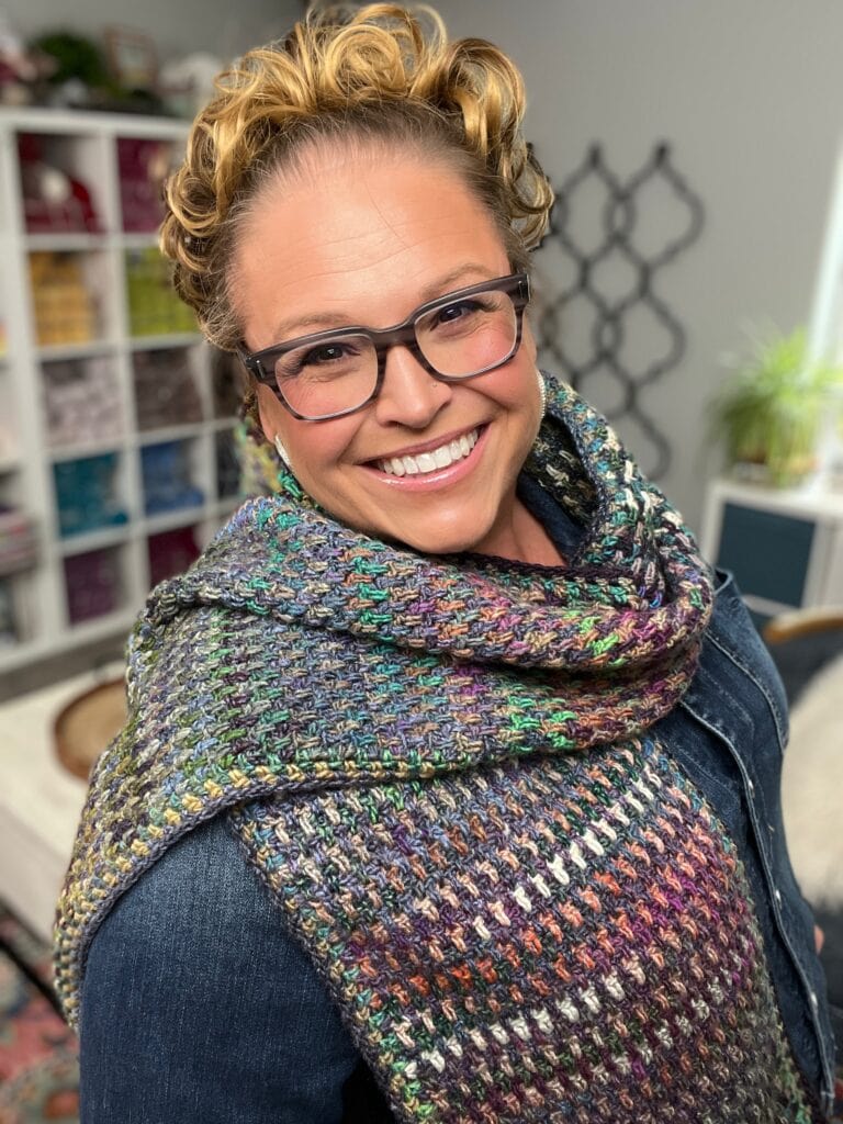 A smiling woman with curly blonde hair, glasses, and a multicolored woven scarf over a denim shirt stands indoors with shelves of yarn and a plant in the background. -Marly Bird