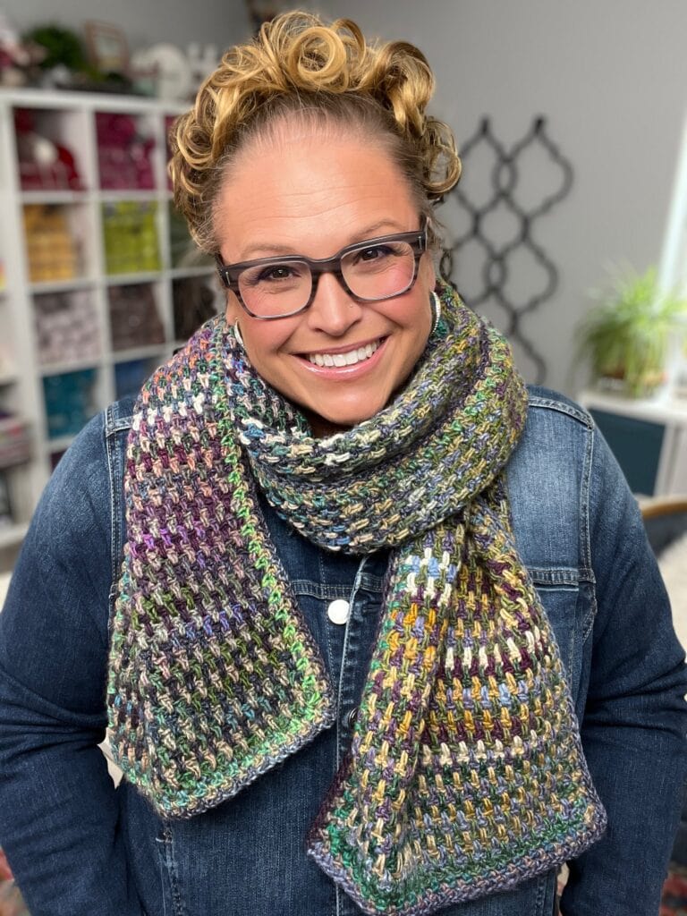 A smiling person with curly blonde hair and glasses wears a textured multicolored knit scarf and a denim jacket, standing indoors with yarn and plants in the background. -Marly Bird