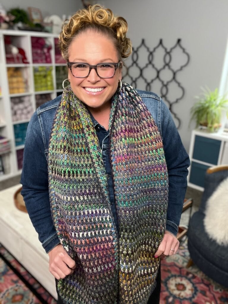 A smiling person with curly hair and glasses wears a colorful, textured scarf over a denim jacket in a cozy, well-lit room with shelves of yarn, a plant, and decorative accents. -Marly Bird