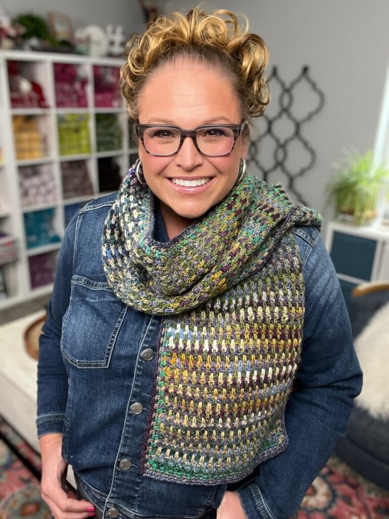 Woman with curly hair and glasses smiling indoors, wearing a blue denim jacket and a multicolored, textured knit scarf. Shelves with yarn and plants are visible in the background. -Marly Bird