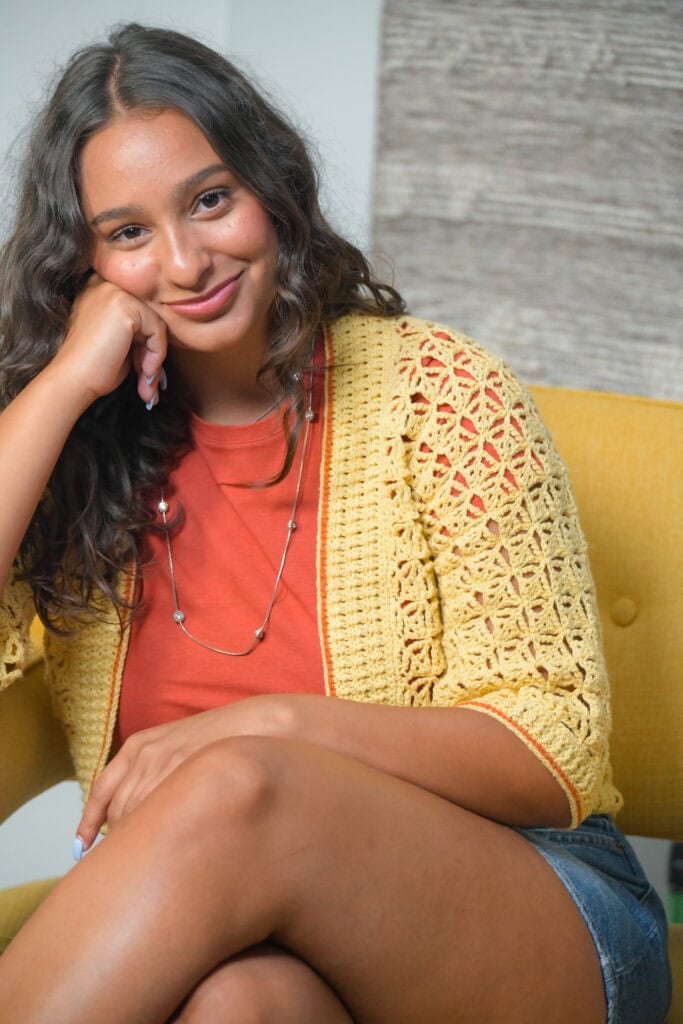 A young woman with long curly hair sits on a yellow chair, smiling with her head resting on her hand. She wears a rust-colored shirt, a yellow crocheted cardigan, denim shorts, and a long necklace. -Marly Bird