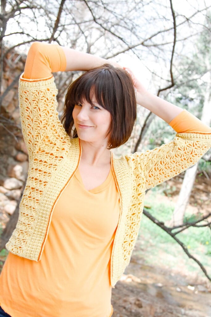 A woman with short brown hair smiles and stretches her arms behind her head. She is wearing an orange shirt and a yellow crocheted cardigan, standing outdoors in front of leafless trees. -Marly Bird