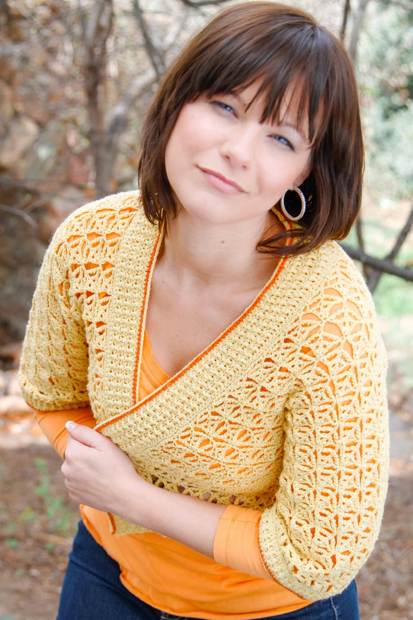 A woman with short brown hair, wearing hoop earrings, an orange top, and a yellow crocheted cardigan, stands outdoors with a soft smile, holding her cardigan closed. The background is blurred with trees and greenery. -Marly Bird