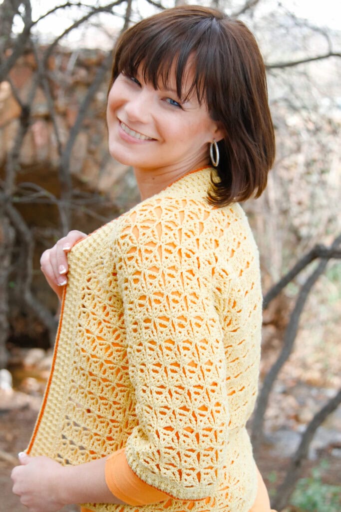 A woman with short brown hair smiles while looking over her shoulder, wearing a yellow crocheted cardigan over an orange top, standing outdoors near trees and branches. -Marly Bird