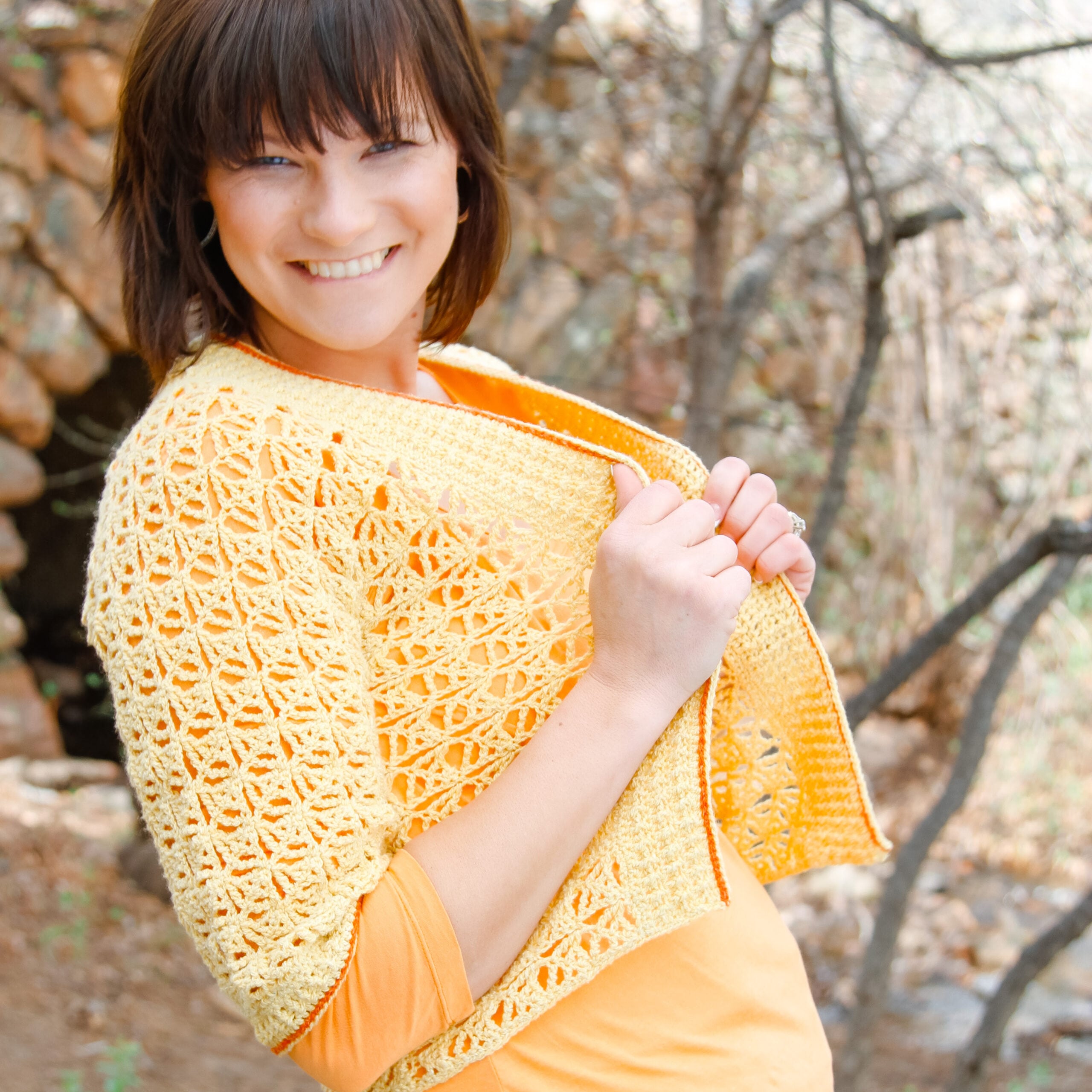 Woman models a goldenrod crochet cardigan with visible textured stitch detail over an orange shirt outdoors near trees and rocks.