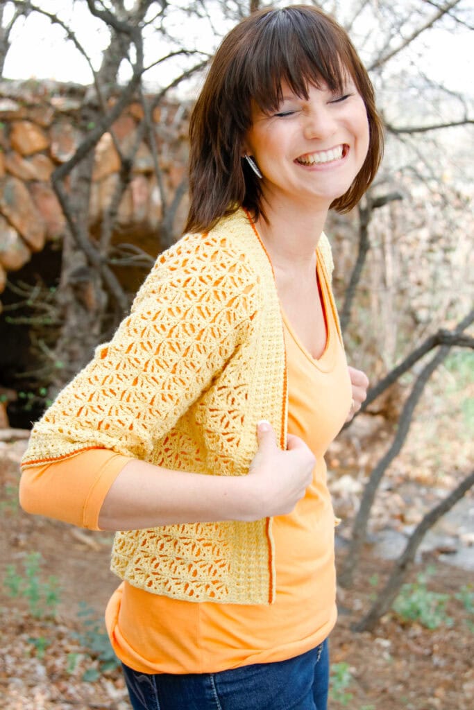 A woman with short brown hair smiles widely outdoors, wearing a yellow crocheted cardigan over an orange top and blue jeans. Leafless trees and rocks are in the background. -Marly Bird