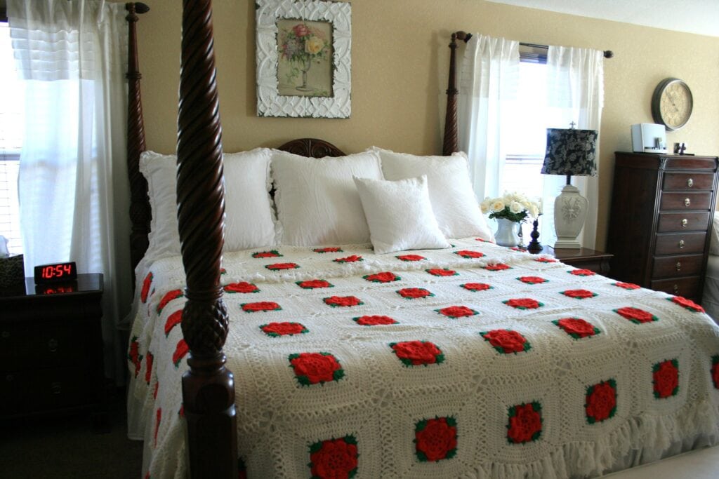 A cozy bedroom features a large bed adorned with a crocheted blanket showcasing intricate flower motifs in red and green. Two windows with white curtains, a framed painting, a wooden dresser, and a digital alarm clock complete the tranquil space. -Marly Bird