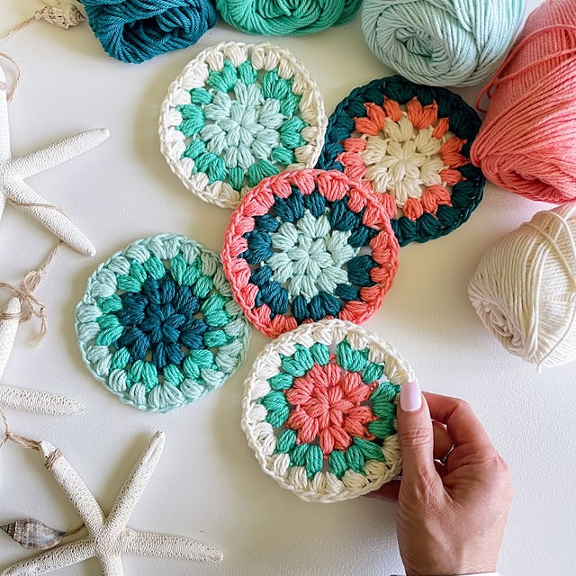 A hand arranges colorful crochet coasters on a white surface, perfect for a touch of spring cleaning charm. The coasters boast circular patterns in teal, coral, and white. Balls of yarn and starfish are scattered around, adding a coastal vibe to the composition. -Marly Bird