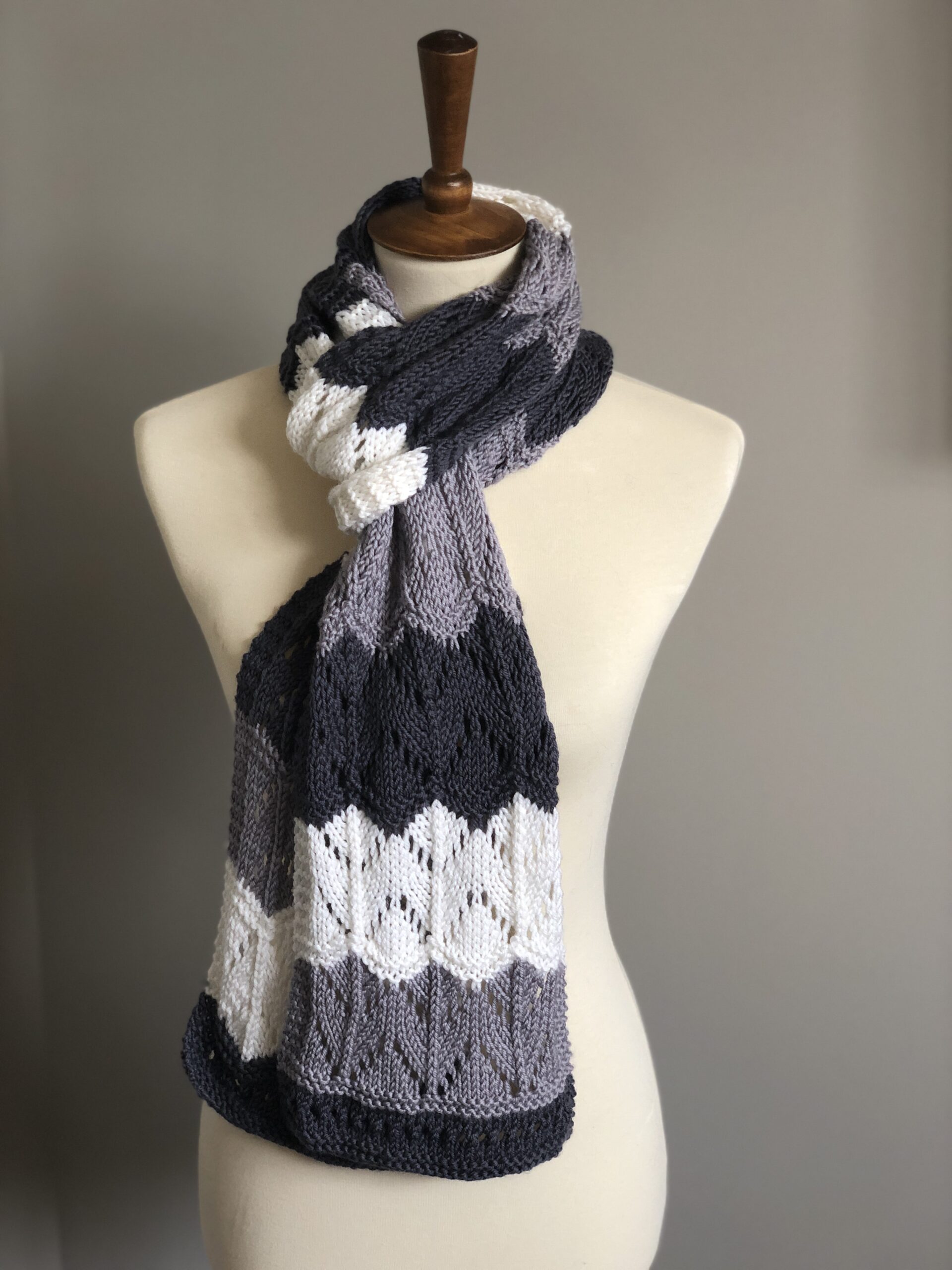 The image displays a mannequin adorned with a beautifully knitted scarf named "Greyson Waves Scarf". The scarf features a striking wave pattern in shades of navy, grey, and white, creating a sophisticated, gradient-like effect. The pattern showcases intricate stitches that give the piece a textured, three-dimensional look. The scarf is elegantly draped around the mannequin's neck, emphasizing its length and the fluidity of the design, reminiscent of the ebb and flow of ocean waves. The color palette and knitting technique suggest a modern twist on classic knitwear, making it a versatile addition to any wardrobe. The overall presentation of the scarf is polished and stylish, indicating a design by Marly Bird, known for chic and contemporary knit patterns.