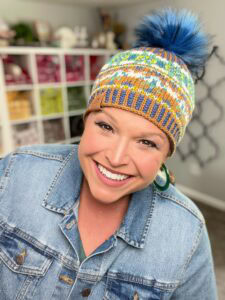 A smiling person wearing a denim jacket and a colorful knit hat with a blue pom-pom stands indoors, surrounded by shelves of yarn—a perfect setting to showcase their mosaic slip stitch knitting pattern. -Marly Bird