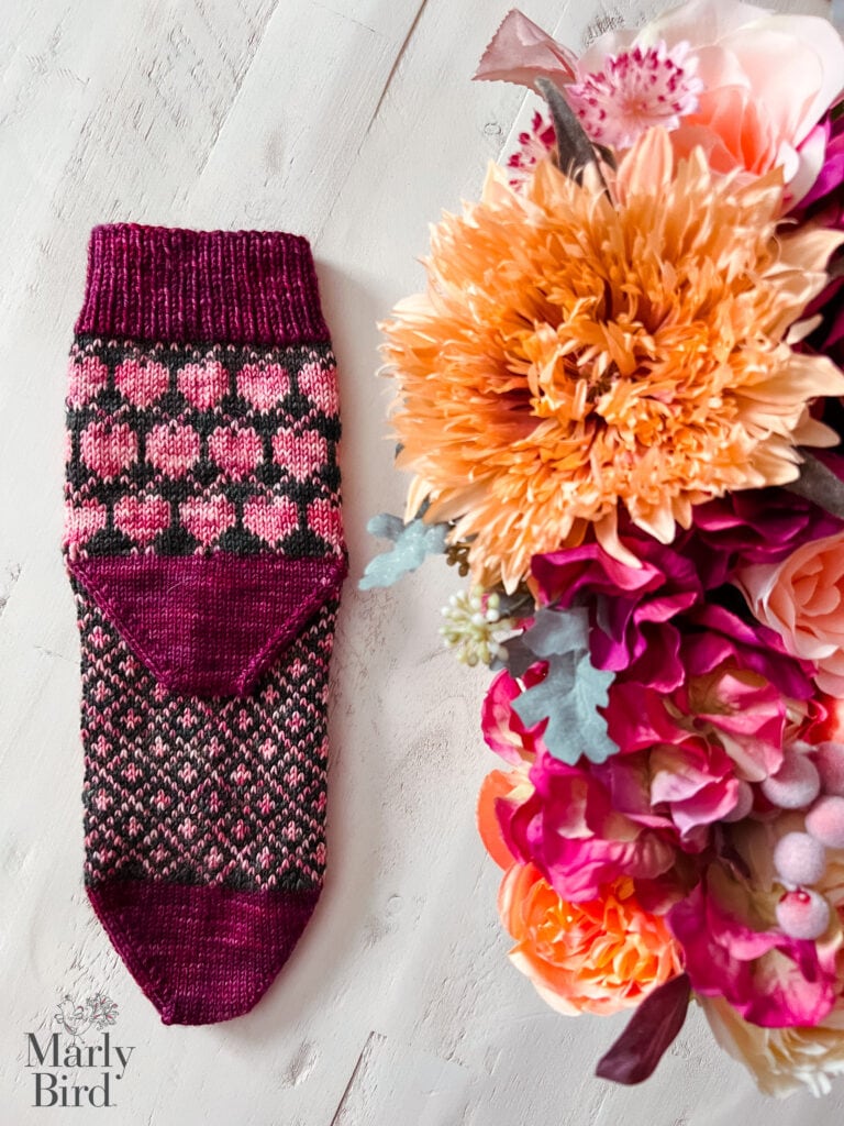 A Heartstrings knit sock pattern with pink and burgundy hearts rests on a white wooden surface beside a vibrant bouquet in orange, pink, and magenta. The "Marly Bird" logo appears in the corner. -Marly Bird