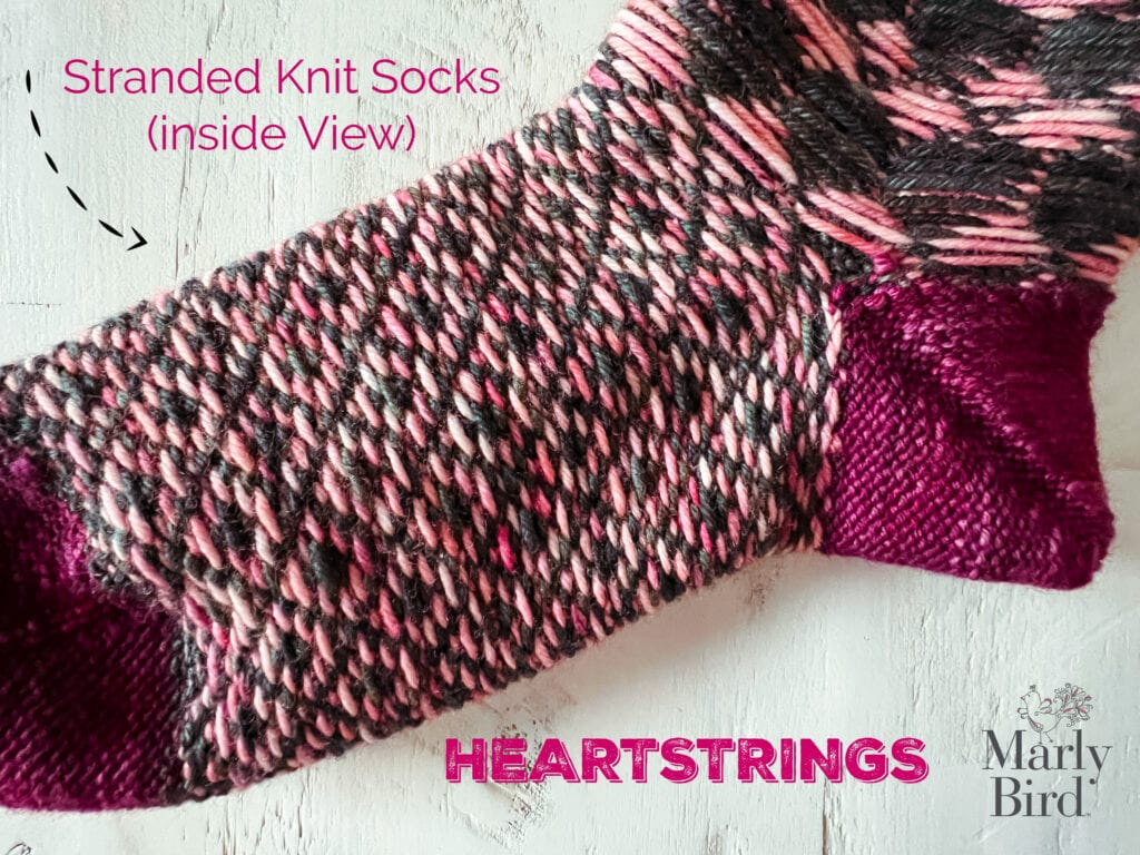 A close-up of the inside view of stranded knit socks in pink, black, and burgundy yarn. Text reads “Stranded Knit Socks (inside view),” “HEARTSTRINGS knit sock pattern,” and “Marly Bird.”. -Marly Bird