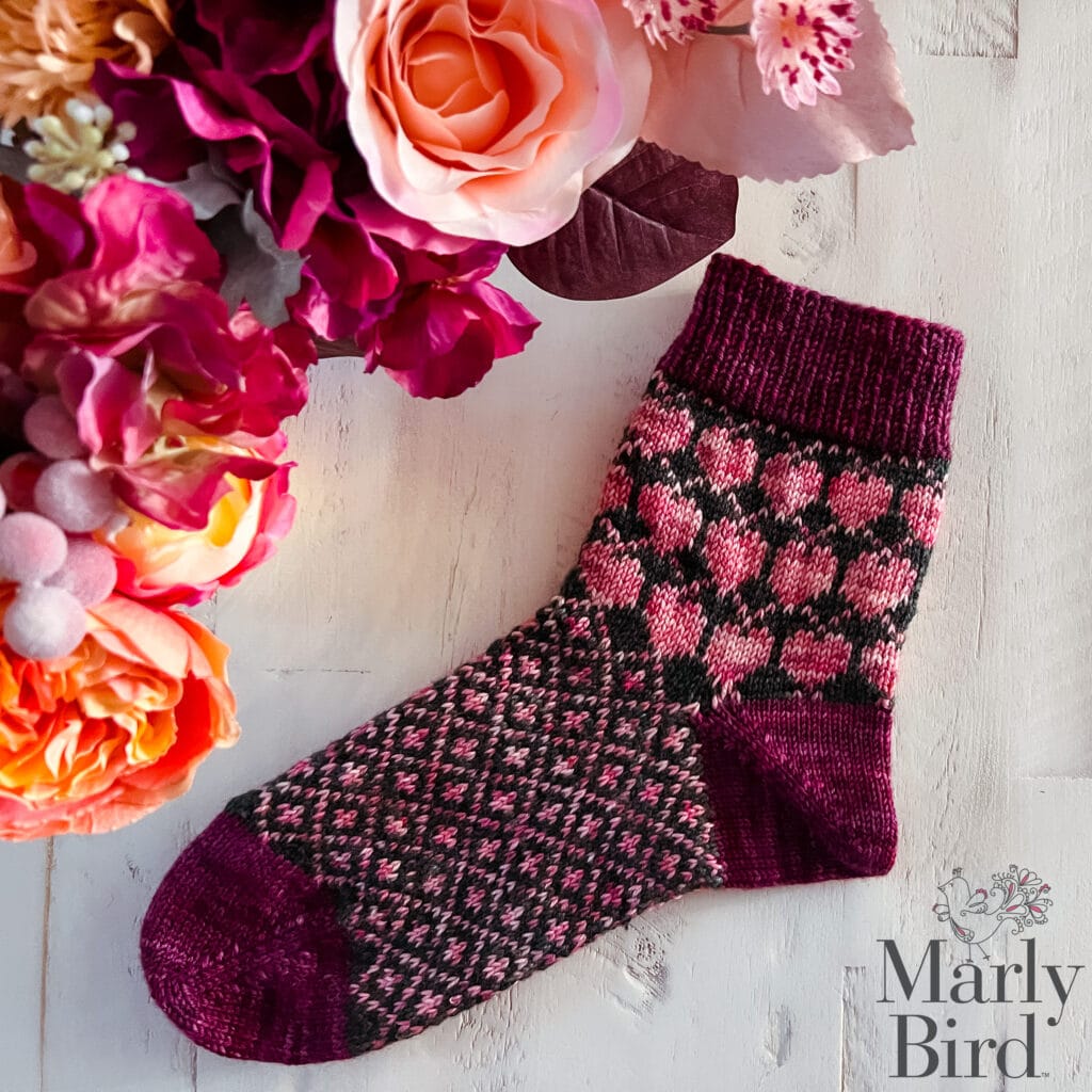A knitted sock with a pink and burgundy heart pattern, made using the Heartstrings knit sock pattern, lies on a white wooden surface next to a vibrant bouquet of pink and orange flowers. "Marly Bird" is printed in the bottom right corner. -Marly Bird