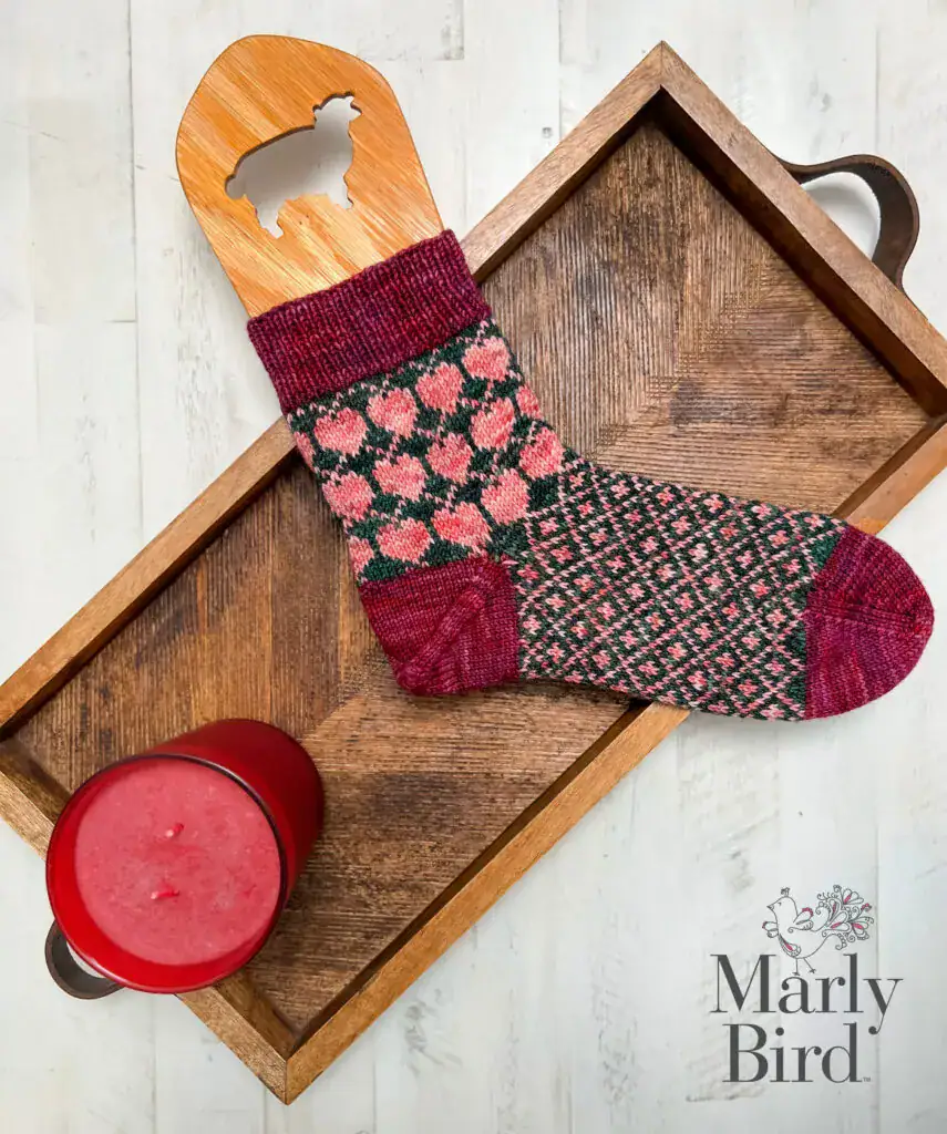 A wooden tray holds a single knit sock with pink and burgundy patterns on a wooden sock blocker, next to a red candle. Discover more free knitting and crochet patterns from Marly Bird—logo visible in the bottom right corner. -Marly Bird