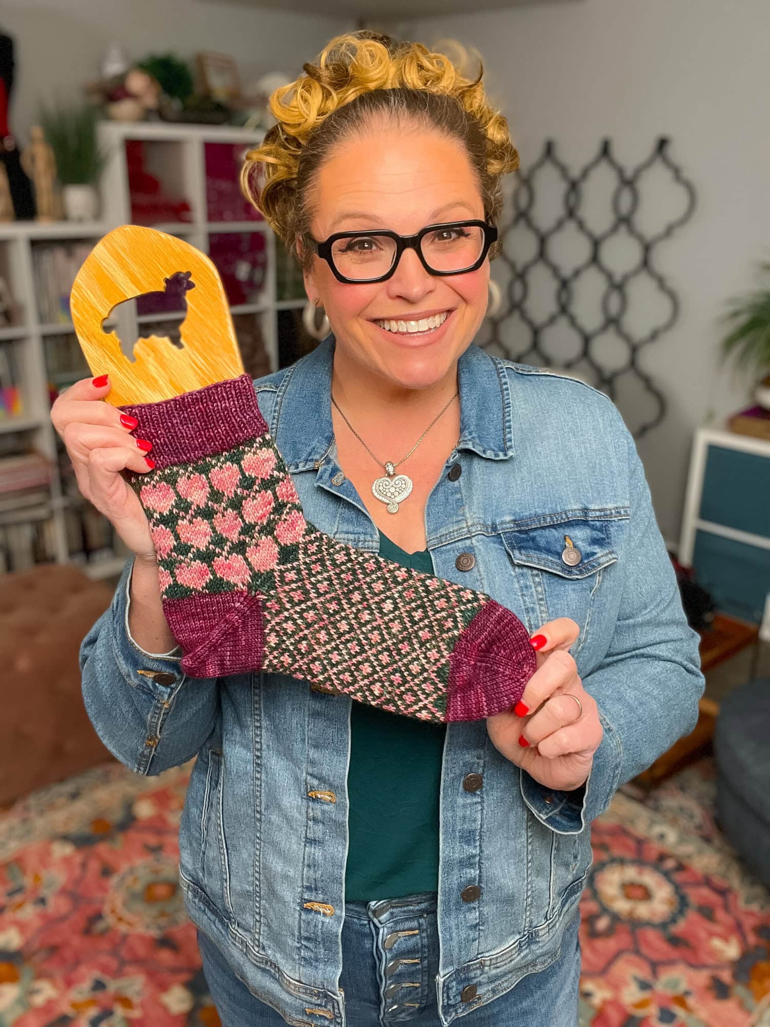 A smiling woman with glasses and curly hair, wearing a denim jacket, holds up a colorful colorwork sock featuring shades of pink, purple, and green. She stands in a cozy, decorated room showcasing her latest knit sock pattern. -Marly Bird