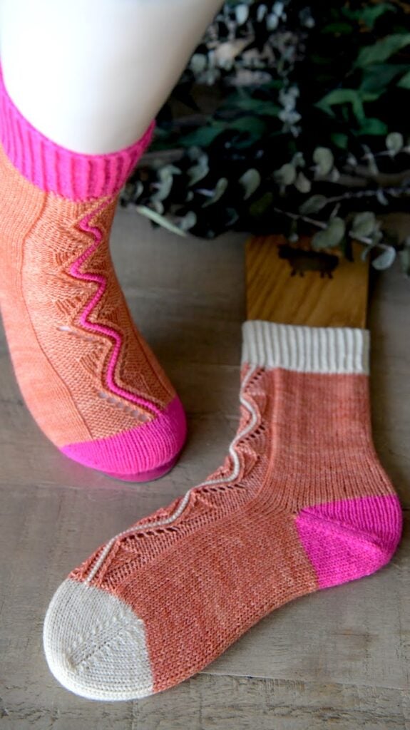 Orange knit socks with wavy pink and white designs; one sock has a pink toe and cuff, the other white. Shown on wood surface.