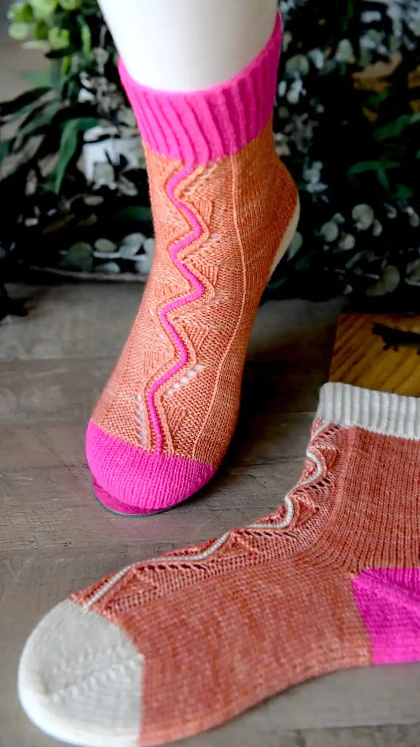 Hand-knit socks with wavy pink stripes on orange; one has white toe/cuff, the other bright pink. Displayed on wood with greenery. Vertical colorwork knit socks using mini skeins