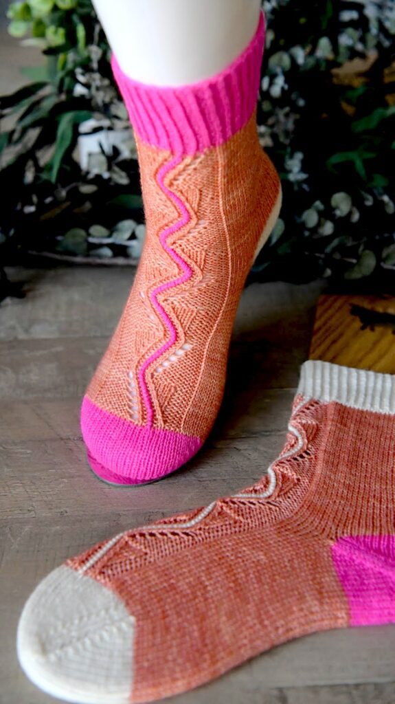 A close-up of two knitted socks displayed on a wooden surface. One sock is being worn and features a pink cuff and toe with an orange and light brown body. The other sock, lying flat beside it, in similar colors but with a white cuff and heel, evokes the charm of Appalachian Cozy Up with its zigzag patterns. -Marly Bird