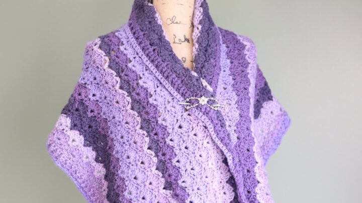Purple Tunisian crochet shawl with a scalloped edge, textured stitches, and a brooch closure on a mannequin.