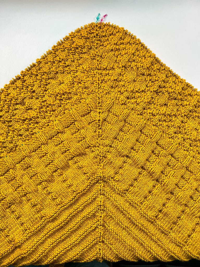 Triangular yellow knit shawl with raised textured stitches, colorful markers at the top, displayed on a white background.