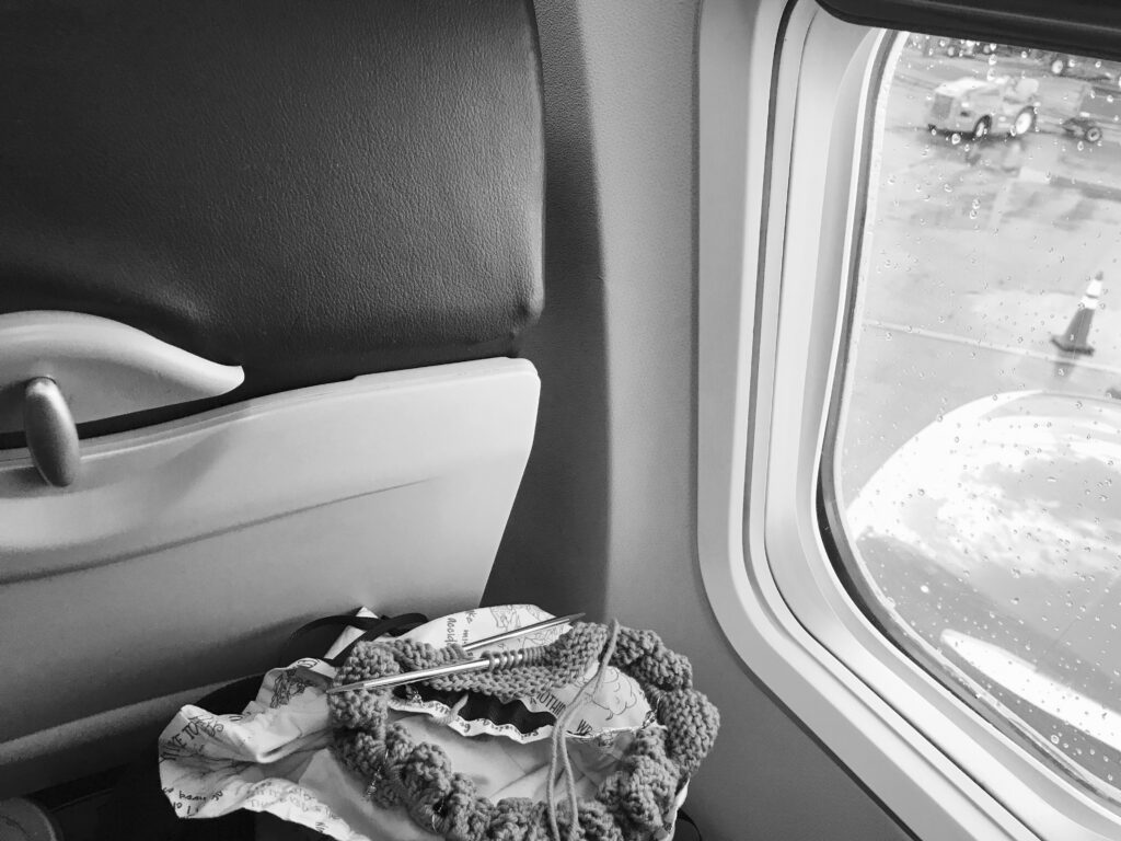 black and white image of a travel knitting project next to the airplane window  before take off - knitting needles on a plane - Marly Bird