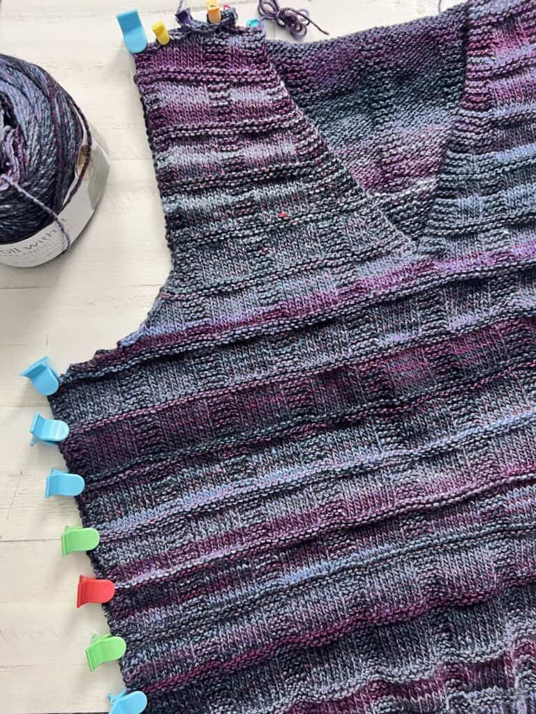 A knitted vest with a purple and gray striped pattern is in progress, featuring colorful clips along the edge. A ball of matching yarn rests nearby on the white surface, ready to add finishing touches for a polished look. -Marly Bird