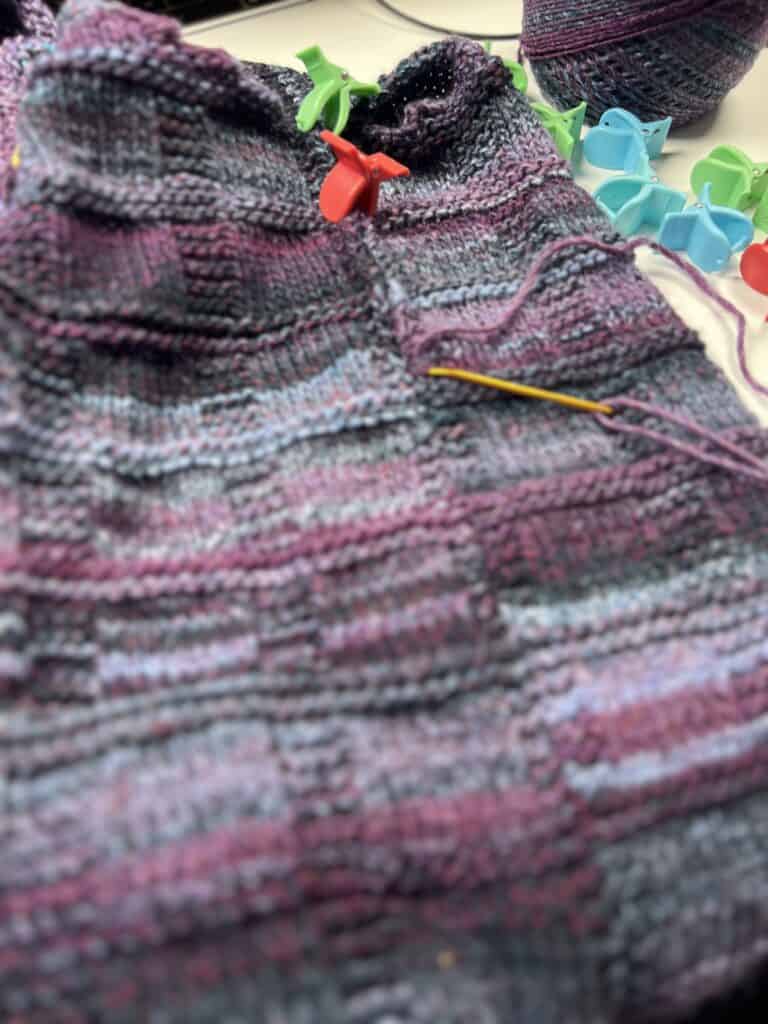 A close-up of a colorful striped knitting project in progress, perfecting the knit vest with purple, blue, and pink hues. Yarn loops and knitting tools, including stitch markers in green and orange, add the finishing touches to this polished look. -Marly Bird