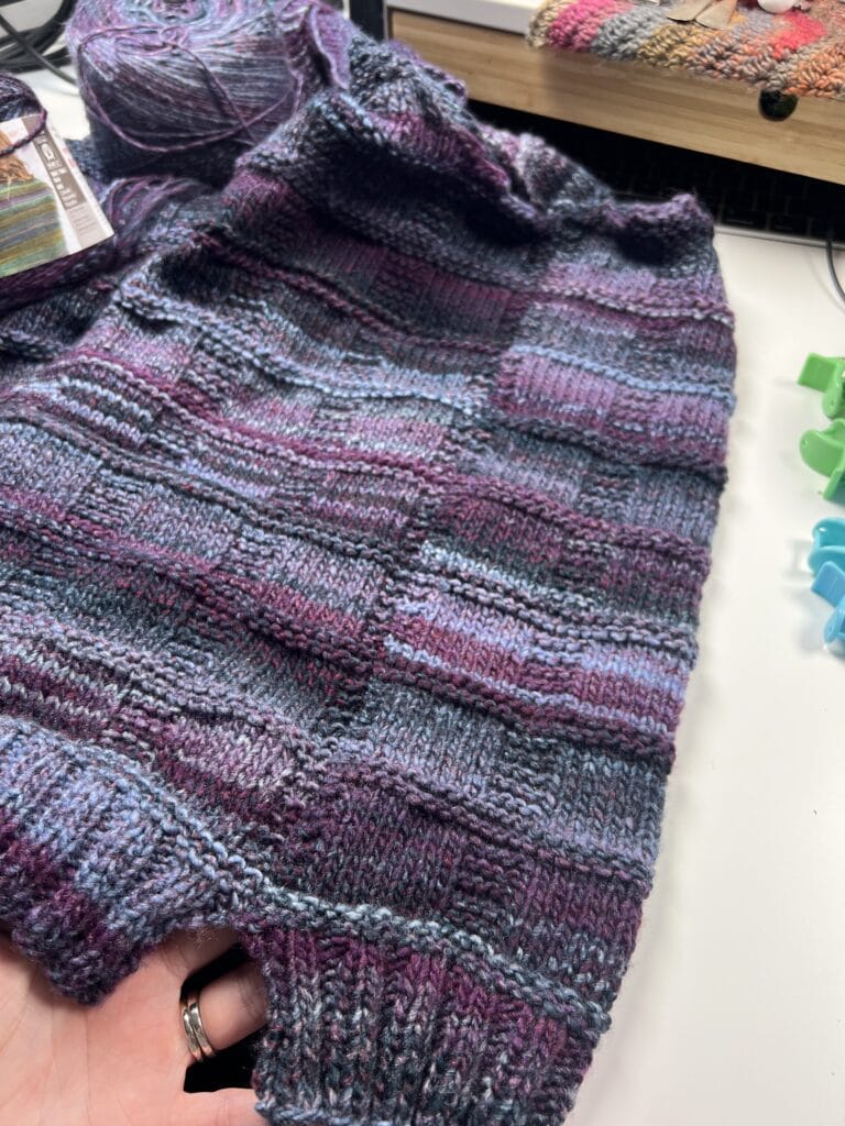 A hand holds a partially knitted Lorelai Knit Vest, crafted with variegated yarn in shades of purple, blue, and burgundy. Knitting supplies and tools are visible in the background on a white surface. Try this free knit vest pattern today!. -Marly Bird