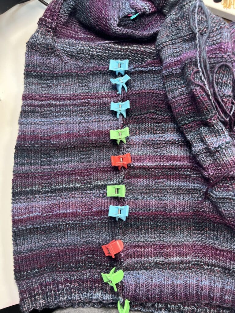 A close-up of a Lorelai Knit Vest, featuring shades of purple, gray, and blue, with colorful plastic stitch markers clipped along the center seam. The matching knitted sleeve hints at this free knit vest pattern’s coordinated style. -Marly Bird