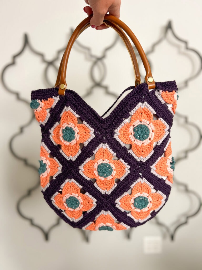 A hand holds a crocheted bag with wooden handles, featuring a pattern of orange, teal, and purple granny squares in a floral design. The background is softly blurred with a geometric wall pattern. -Marly Bird