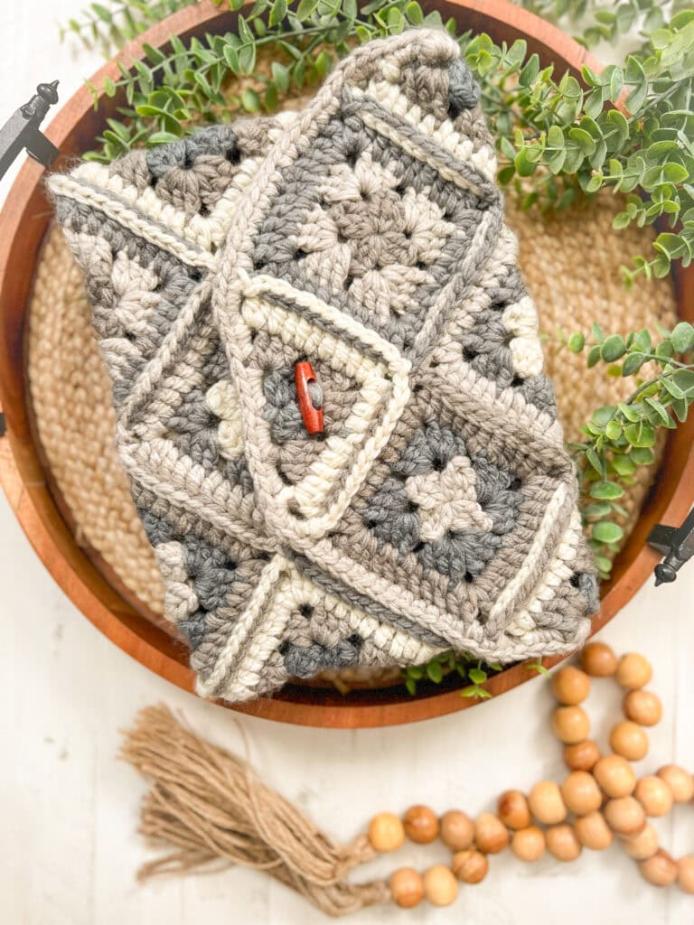 A crocheted clutch made with a One Ball Crochet Granny Square Pattern in neutral tones rests in a wooden bowl, surrounded by greenery, woven mat, and wooden beads with a tassel. A small red button secures the flap. -Marly Bird