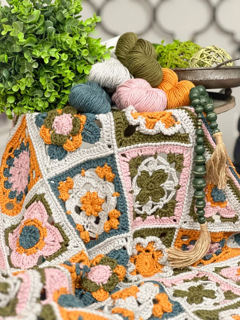A colorful crochet blanket with floral granny squares in pink, orange, green, and white is draped over a surface, surrounded by yarn balls, a green beaded tassel, and two potted plants. -Marly Bird