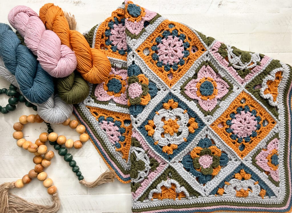 Colorful granny square crochet blanket with floral patterns in pink, mustard, green, and gray, displayed next to skeins of yarn in matching shades and a wooden bead garland with tassels on a light surface. -Marly Bird