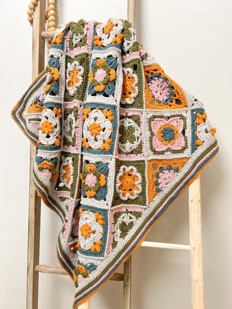 A colorful crocheted blanket with various square patterns in green, orange, pink, and white draped over a wooden ladder against a plain light wall. -Marly Bird