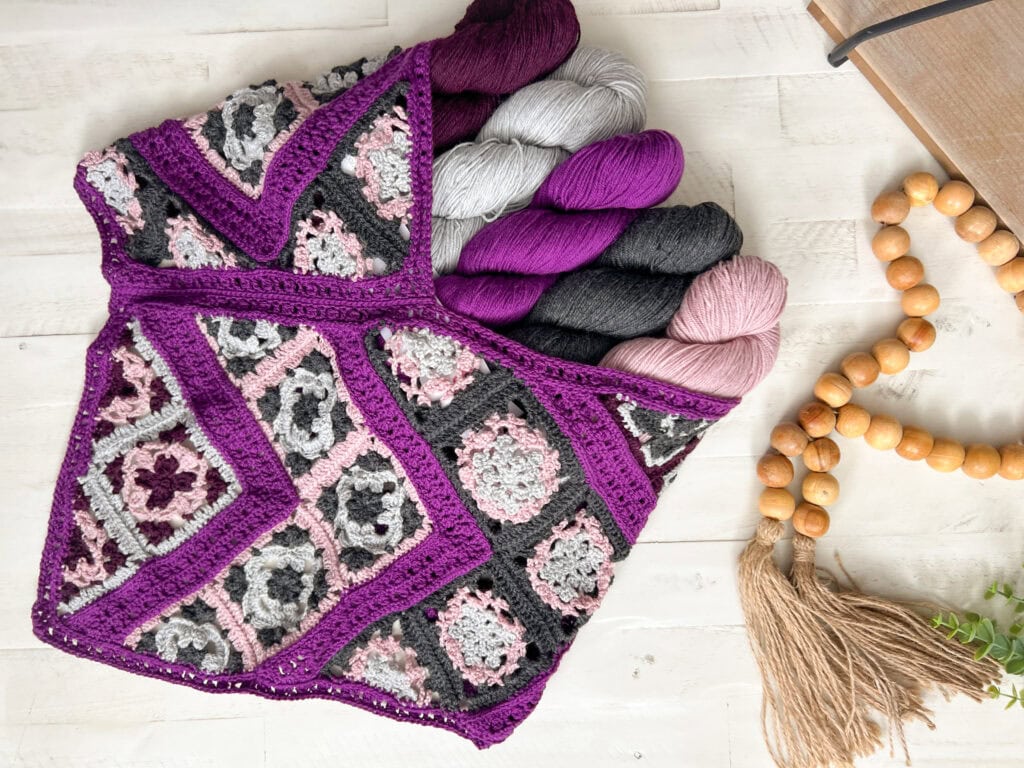 A crocheted bag with purple, grey, pink, and black granny squares holds skeins of yarn in matching colors. The bag rests on a white wooden surface next to wooden prayer beads with a tassel. -Marly Bird