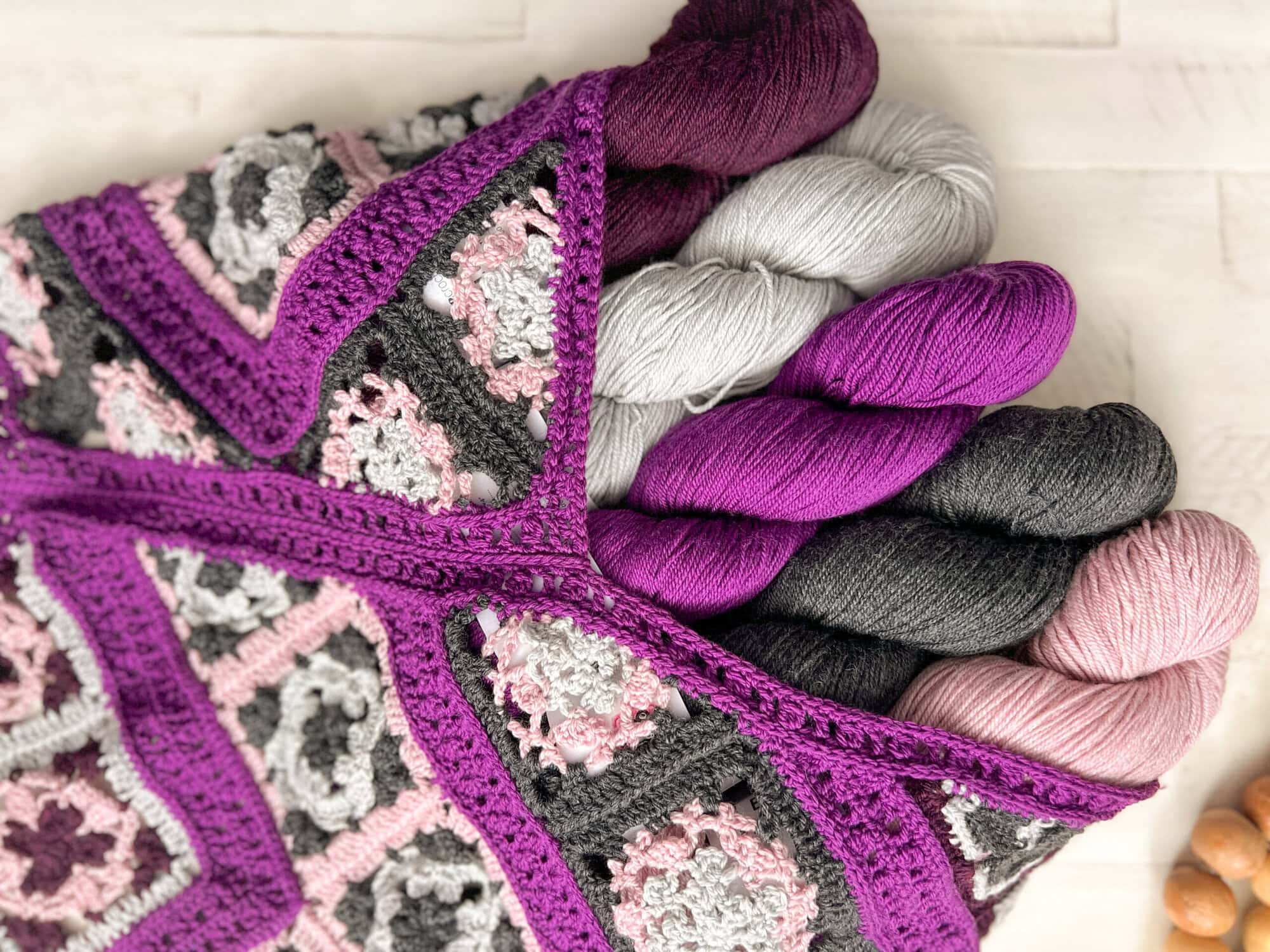 A crocheted bag with a geometric pattern in purple, gray, pink, and white holds five skeins of yarn in shades of purple, gray, and pink on a light wooden surface. -Marly Bird