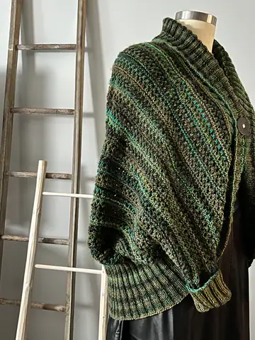 A mannequin displays a textured, green and brown crocheted shrug with ribbed cuffs and a large button—perfect inspiration for free knitting and crochet patterns—positioned beside two wooden ladders against a light gray wall. -Marly Bird