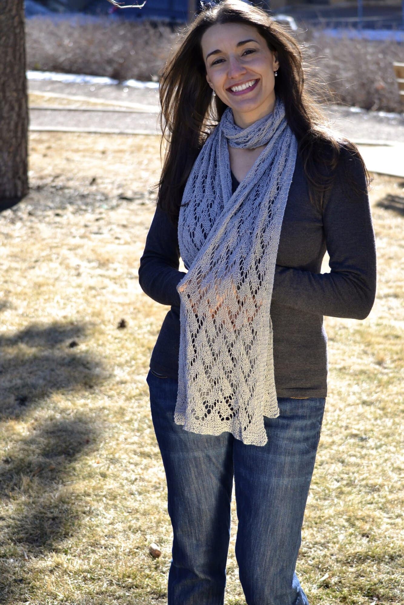 Marly Bird laughing and holding the ends of the Shangri-La free knit lace shawl scarf pattern in soft gray.
