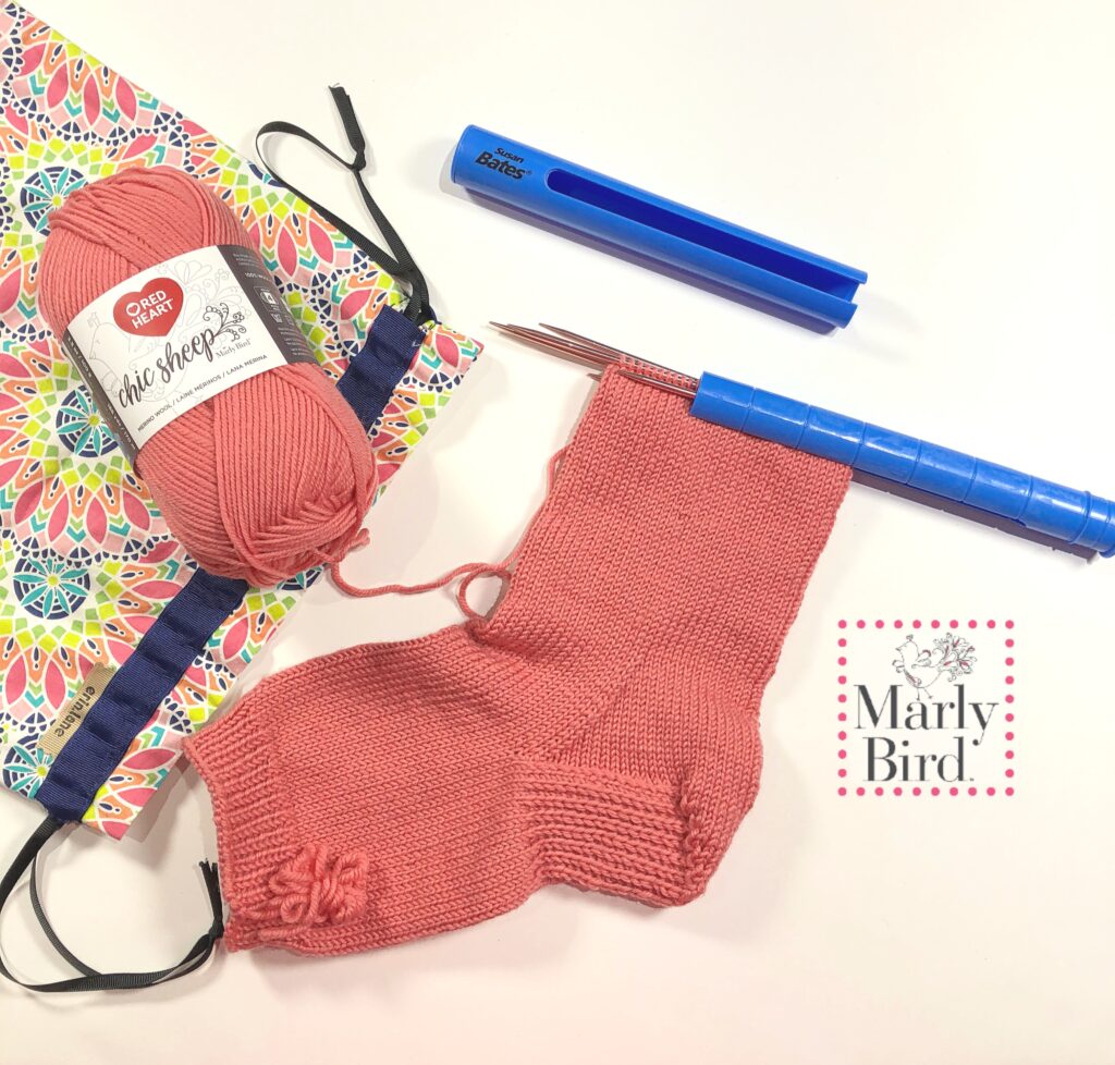 A partially knitted pink sock takes center stage, accompanied by a matching yarn ball labeled "the sheep," a colorful patterned notebook, and blue knitting needle holders—perfect for those who love to create. Celebrate your craft with the Marly Bird logo proudly displayed on the right. -Marly Bird