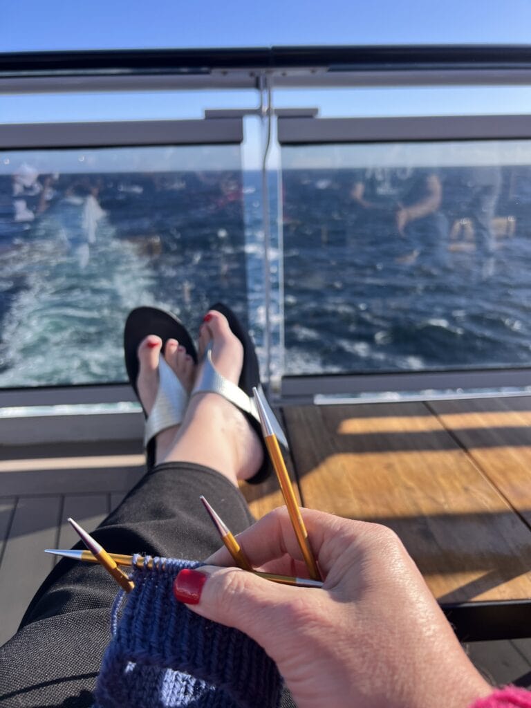 Close-up of hands with red nail polish knitting with wooden needles by the Douro River; fine stitches and yarn texture visible.