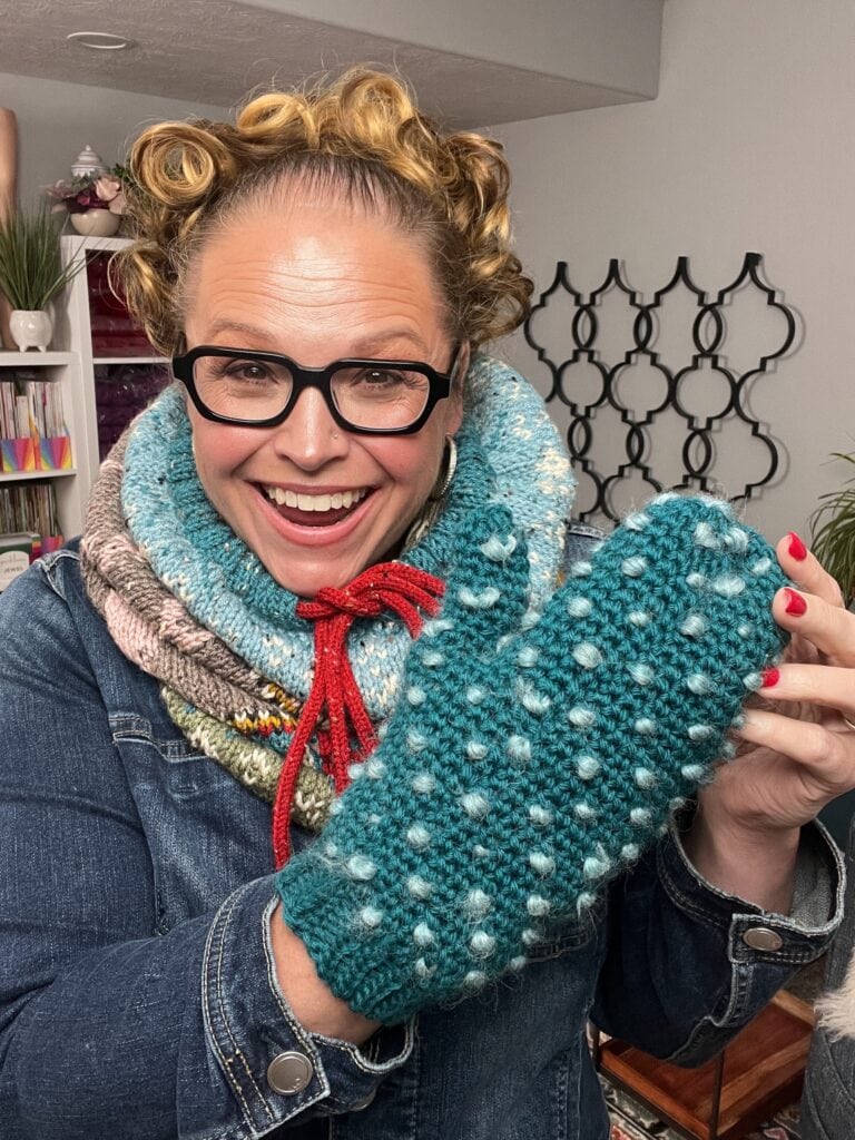 A smiling woman with curly hair, black glasses, and a colorful knitted scarf poses indoors, holding up a teal knit mitten with white bobbles—an Andes-inspired accessory she made using a free knitting pattern. -Marly Bird